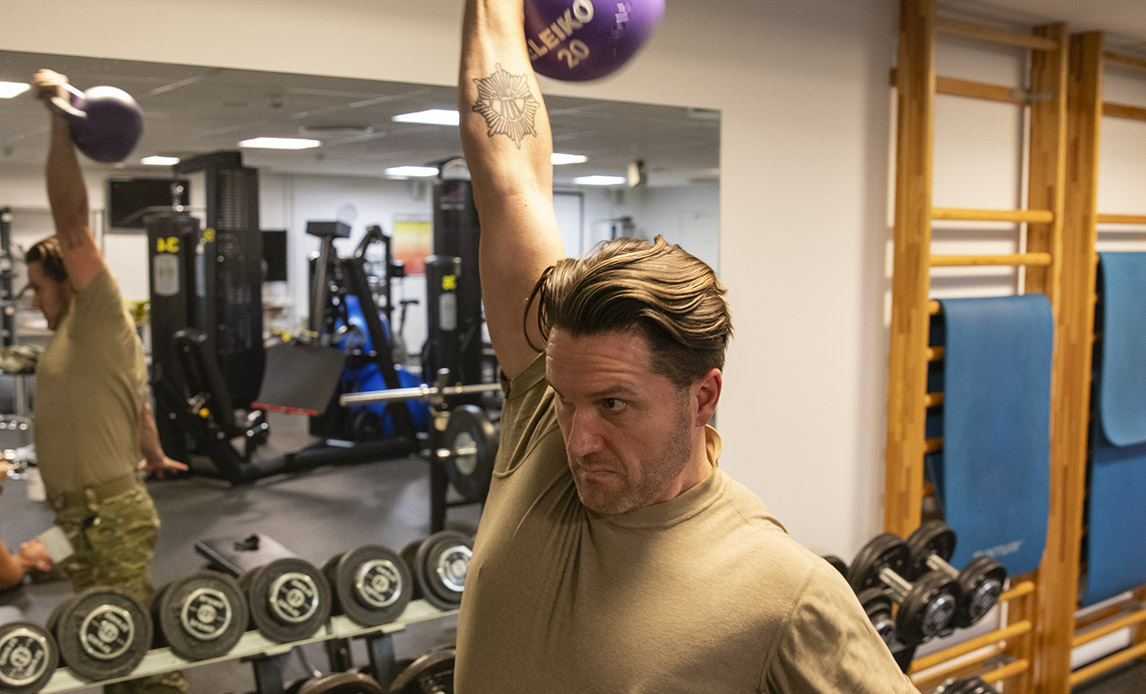 En soldat i uniform i et træningscenter. Han løfter en kettlebell i strakt arm over sit hoved